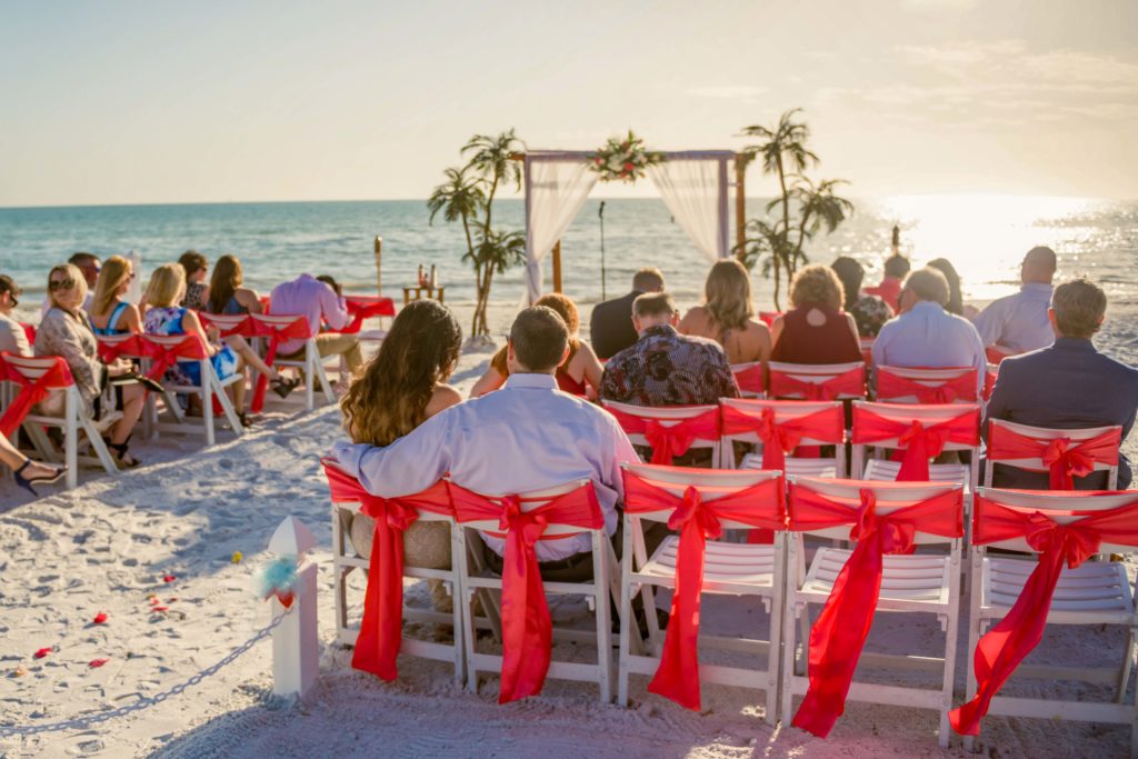 Four rows of chairs with red sashes tied in a bow. Background is bamboo arch and shoreline.