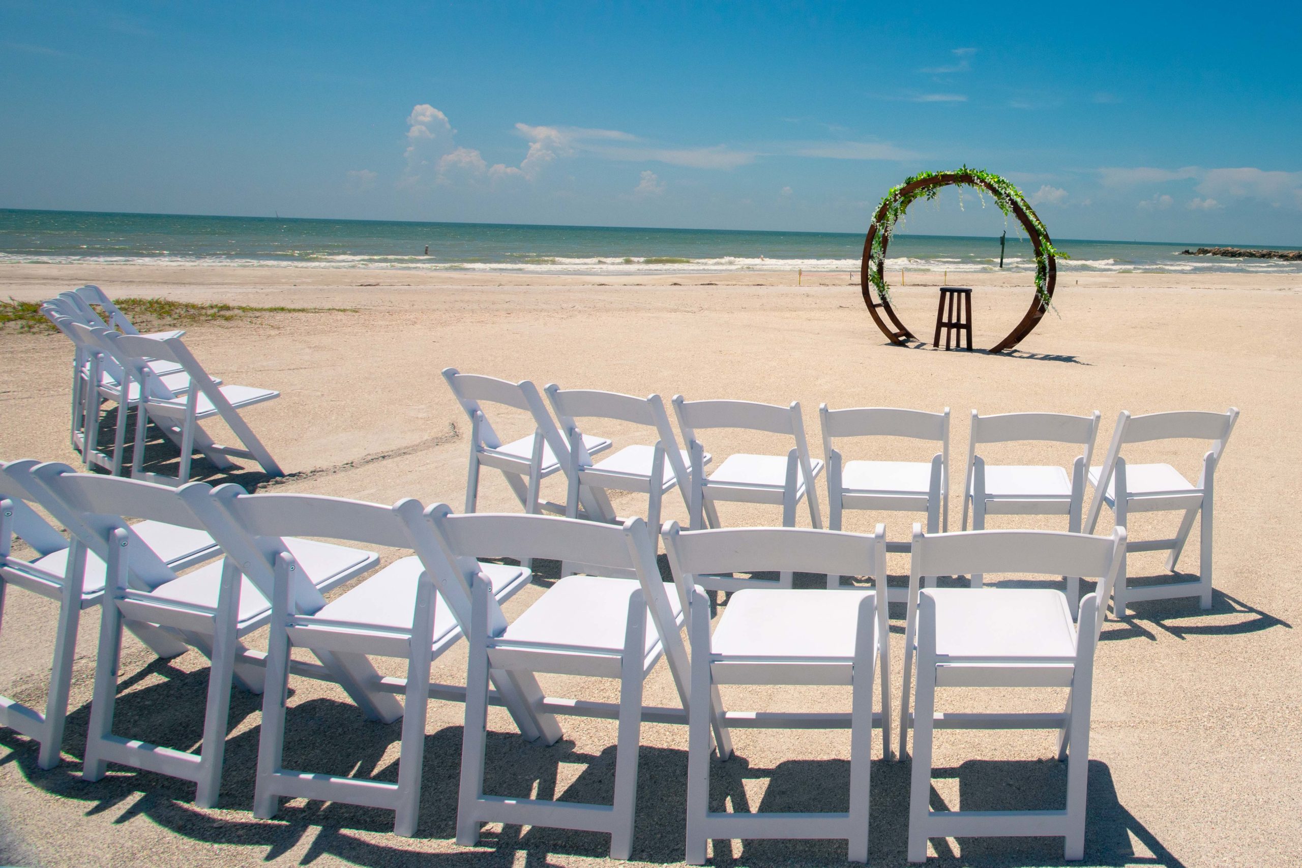 Two rows of chairs arranged in a semi-circle before circular wooden arch on the beach.