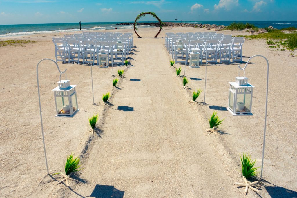 Aisle lined with seagrass and star fish, in addition to small white lanterns filled with seashells and candles hanging from sheppards' hooks. Aisle leads to a circular wooden arch in front of gulf.