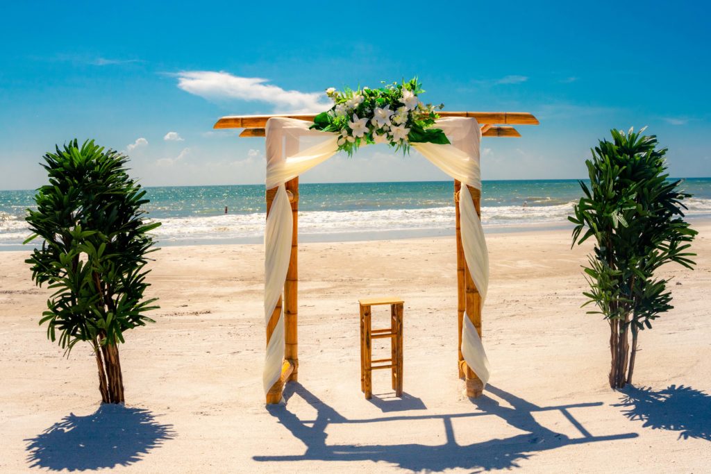 A four-post bamboo arch draped in white silk and topped with white flowers. On either side are tall palm trees, and the Gulf is in the background.