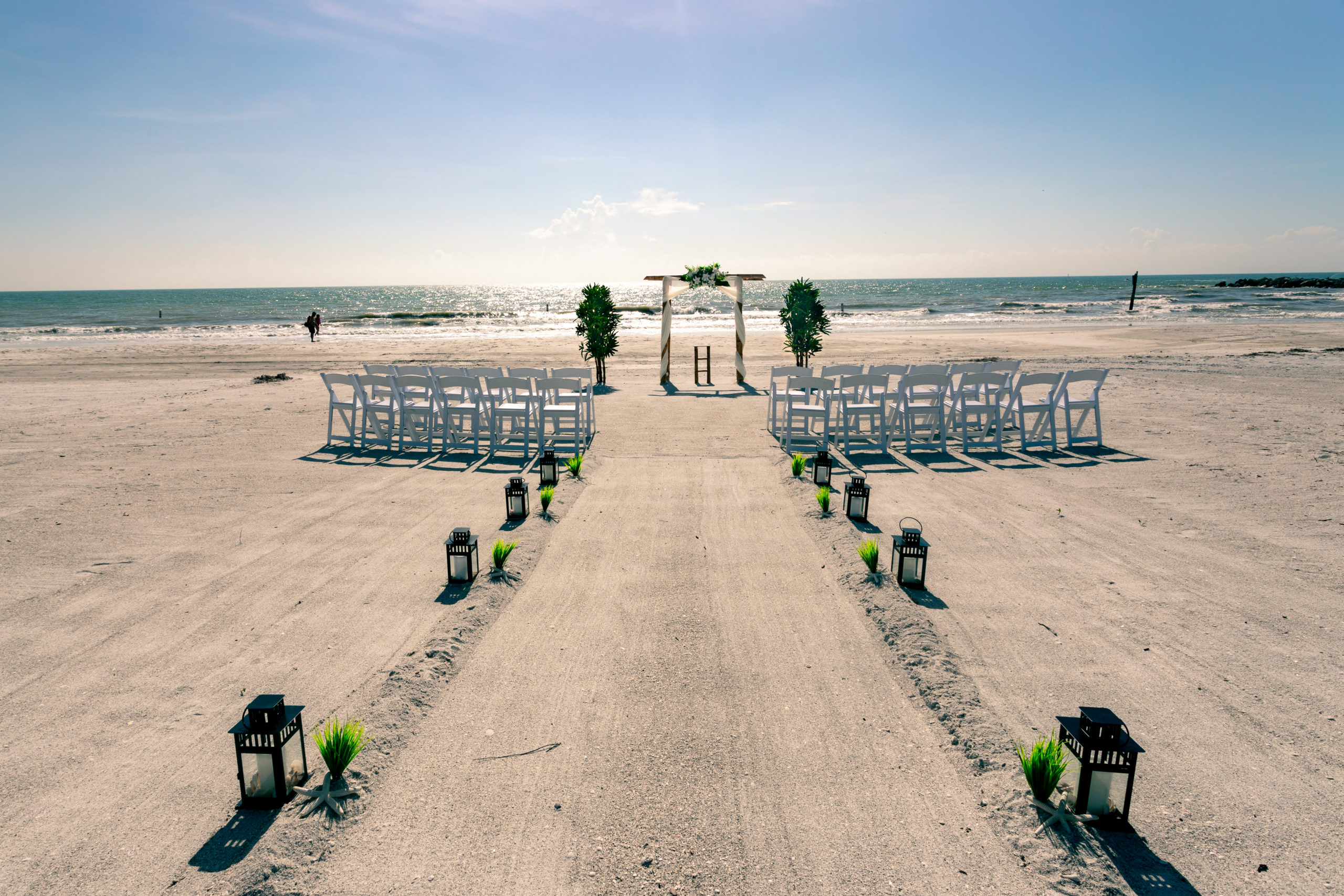 Small black lanterns line a raised aisle of sand leading to an arch on the beach.