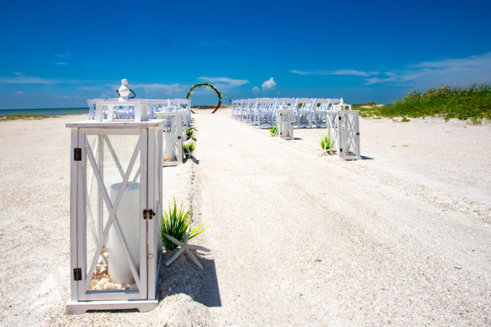 Large white lanterns line an aisle in the sand leading to a circular arch.